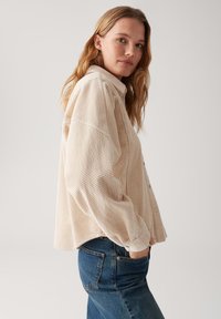 Woman with long blonde hair wearing a loose beige corduroy button-up shirt and blue jeans with hands in pockets, facing sideways.