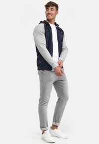 Navy and gray zip-up hoodie with a hood, featuring beige accents and a logo. Paired with light gray cuffed jeans and white sneakers.