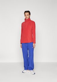 Maglia rossa in pile a maniche lunghe con colletto alto e zip frontale; abbinata a pantaloni blu larghi e impermeabili; scarpe da ginnastica bianche semplici.