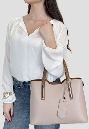 Mujer con blusa blanca y jeans azules sostiene un bolso beige con asas marrones sobre un fondo liso.