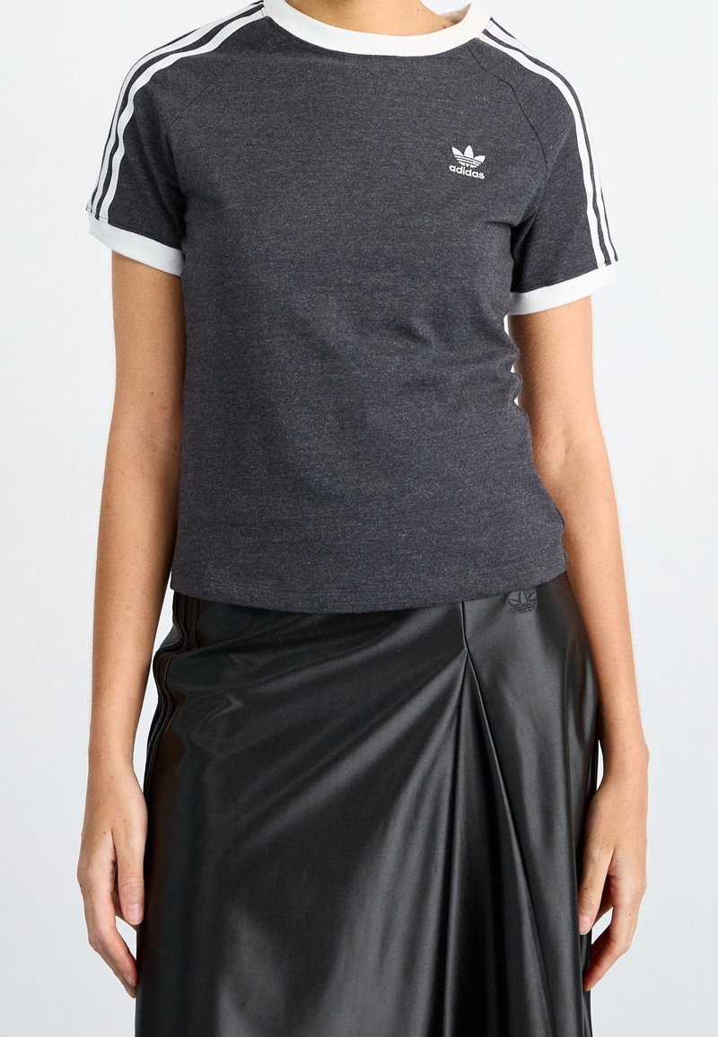 Žena v tmavě šedém tričku Adidas s bílými pruhy na rukávech a černé plisované kožené sukni.