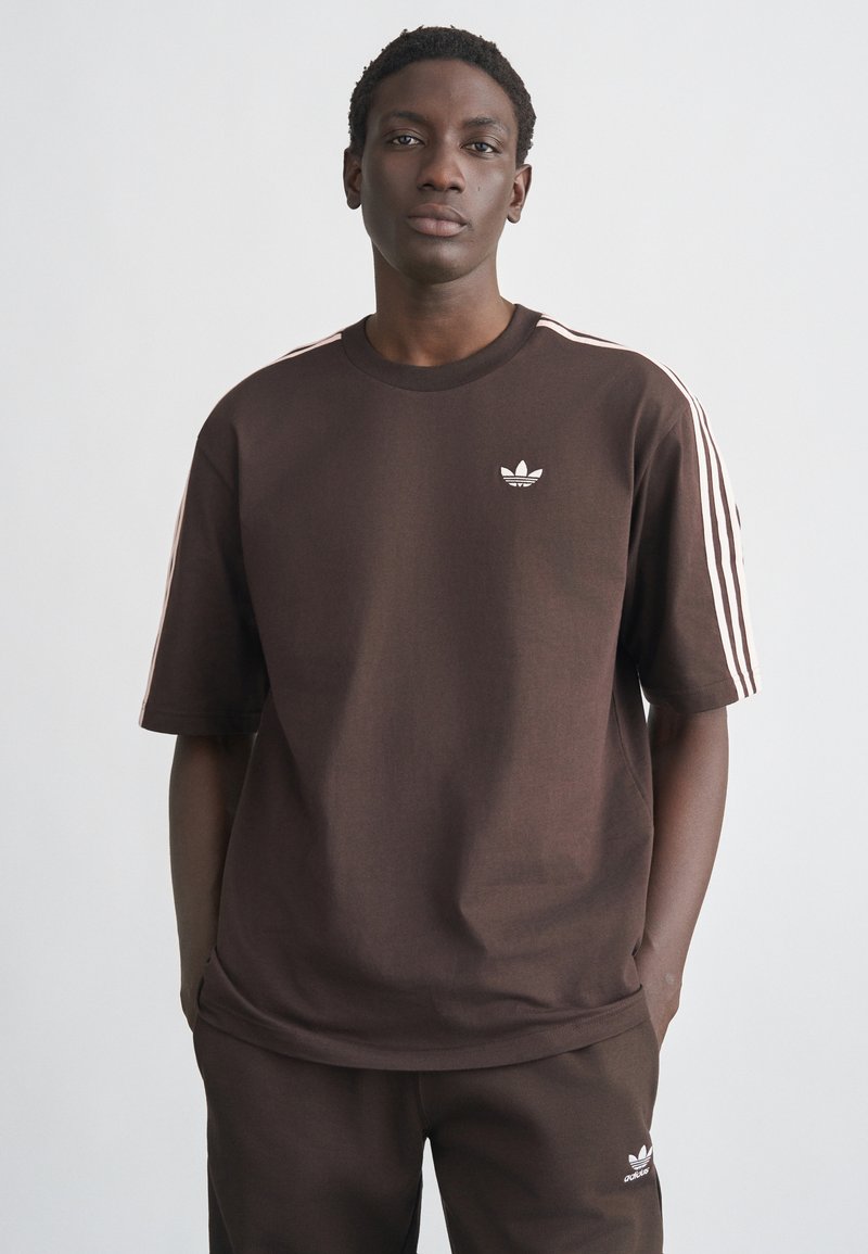 Καφέ βαμβακερό μπλουζάκι με κοντά μανίκια, με λευκό λογότυπο Adidas στο στήθος και λευκές ρίγες στους ώμους.