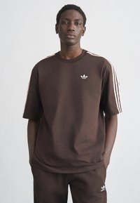 Brown cotton t-shirt with short sleeves, featuring white Adidas logo on the chest and white stripe accents on the shoulders.