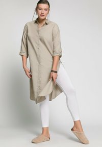 Robe chemise beige à boutons avec des manches retroussées, associée à des leggings blancs et des chaussons en fourrure beige. Texture lisse et coupe décontractée.