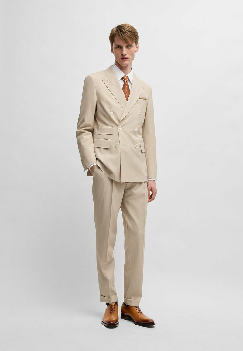Beige double-breasted suit with notch lapels, three pockets, and five buttons. Paired with a white shirt, brown polka dot tie, and brown leather shoes.