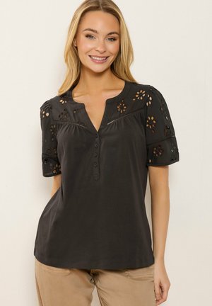 Frau trägt eine schwarze Kurzarmbluse mit floralen Ausschnitten und Knöpfen, kombiniert mit beigen Hosen, lächelt vor einem einfarbigen Hintergrund.