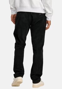 Black corduroy pants with a smooth texture, two back pockets with buttons, and a straight leg fit. Worn with a light gray sweatshirt and sneakers.