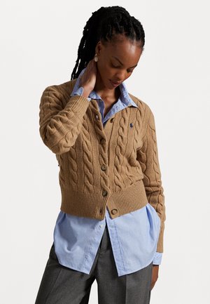 Frau mit geflochtenem Haar trägt eine beige, grob gestrickte Strickjacke über einem blauen Button-up-Hemd und grauen Hosen und steht mit einer Hand an ihrem Hals.