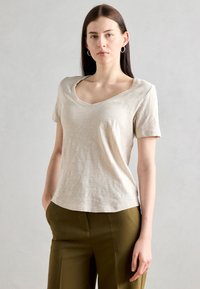 Tricou bej cu decolteu în V, dintr-un material moale, cu mâneci scurte, croială lejeră, purtat cu pantaloni verzi măslini, design simplu, textură netedă.