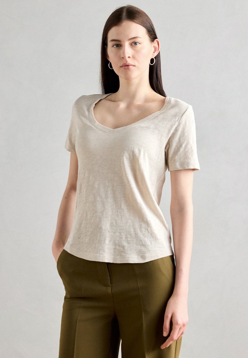 Tricou bej cu decolteu în V, dintr-un material moale, cu mâneci scurte, croială lejeră, purtat cu pantaloni verzi măslini, design simplu, textură netedă.