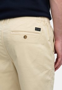 Shorts en coton beige avec une poche arrière à fermeture à bouton et une petite étiquette noire avec "NINETEEN NINETY THREE" en texte blanc.