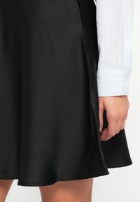 Crna saten suknja s glatkom teksturom, suknja u obliku zvona, s rubom koji završava iznad koljena. Vidljiv je rukav svijetloplave prugaste košulje.