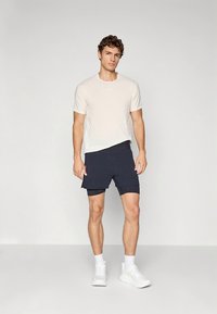 Vaaleabeigea pyöreäkauluksinen t-paita yhdistettynä tummansinisiin shortseihin. Malli käyttää valkoisia urheilukenkiä ja esittelee rentoa urheilullista tyyliä.