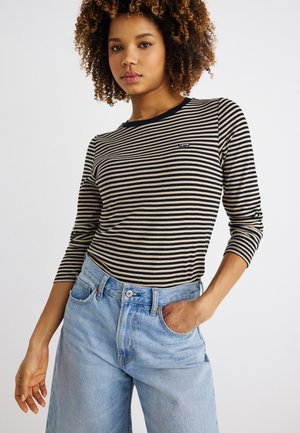 Γυναίκα με κατσαρά μαλλιά φοράει μακρυμάνικη μπλούζα με μαύρες και μπεζ ρίγες και ανοιχτόχρωμο τζιν ψηλόμεσο, στέκεται με το ένα χέρι στην τσέπη.