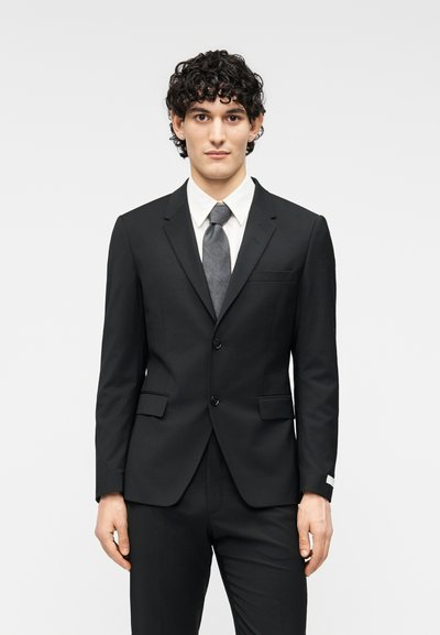 Μαύρο κοστούμι με πέτο με εγκοπή, δύο κουμπιά και πλαϊνές τσέπες. Συνδυάζεται με λευκό πουκάμισο και γκρι γραβάτα, με σχεδίαση στενής εφαρμογής.