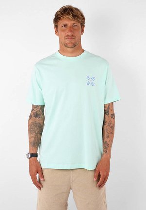 T-shirt vert menthe à manches courtes en coton avec un col rond. Présente un petit motif floral bleu sur la zone à gauche de la poitrine.