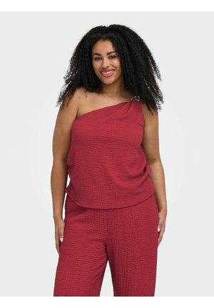 Donna con capelli lunghi e ricci che indossa un top rosso testurizzato monospalla e pantaloni abbinati, sorridente con le mani ai lati.