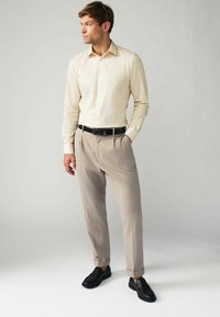 Chemise beige clair à manches longues, pantalons gris clair plissés, ceinture noire et mocassins noirs, se tenant devant un fond neutre.