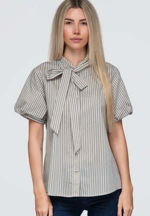 Jonge vrouw met steil blond haar, die een gestreept kortemouwenblouse draagt met een grote strik bij de hals.