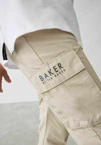 Beige cargo-housut, joissa on strukturoidun pinnan viimeistely, ja isossa sivupussissa on etiketti "BAKER by TED BAKER".