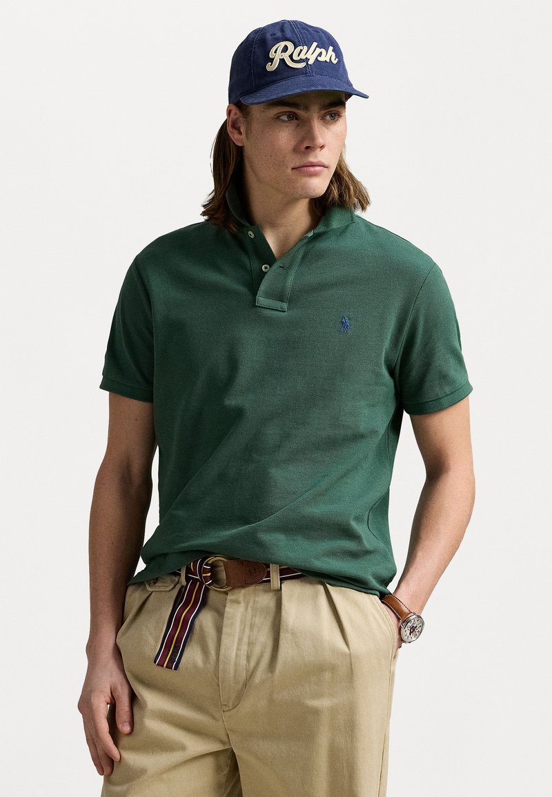 Groene poloshirt met een twee-knopssluiting en korte mouwen, gecombineerd met kaki broek en een blauwe pet met de tekst "Ralph".