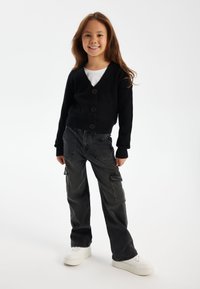 Schwarze gerippte Strickjacke mit drei Knöpfen, kombiniert mit dunkelgrauen Cargo-Hosen mit Seitentaschen und weißen Sneakern.