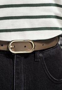 Brun läderbälte med en gyllene oval spänne, bärs över mörka denimjeans, med en vit och grön randig topp synlig ovanför.