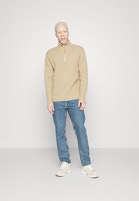 Maglione beige a costine con zip a metà, jeans blu casual e scarpe da ginnastica chiare. Il maglione ha un colletto alto e una vestibilità rilassata.