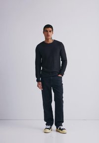 Mand står op ad en ensfarvet væg iført sort langærmet skjorte, mørke jeans og sort-hvide sneakers, med tatoveringer synlige på halsen og ansigtet.