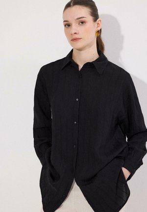 Femme aux cheveux bruns attachés, portant des boucles d'oreilles ovales ambrées et une chemise noire à manches longues boutonnée, les mains dans les poches, sur fond blanc.