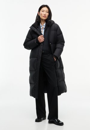 Femme portant un long manteau matelassé noir par-dessus un pull foncé, une chemise bleu clair, un pantalon large noir et des chaussures noires, debout devant un fond blanc uni.