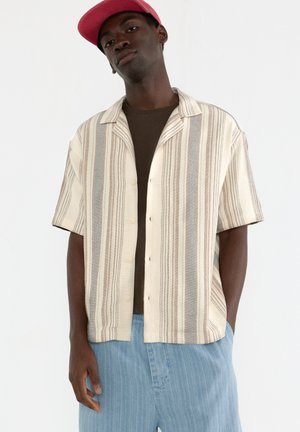 Short-sleeve, beige and gray striped shirt with buttons, paired with a brown shirt underneath and blue striped shorts. Model wearing a pink cap.