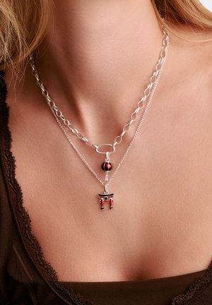 Due collane d'argento al collo di una persona, una con un ciondolo lanterna rossa, l'altra con un ciondolo porta torii rosso e nero.