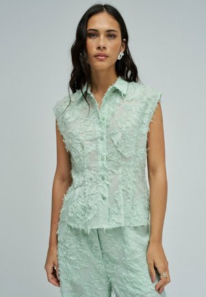 Mujer con el cabello largo y oscuro, que lleva un top sin mangas con botones, textura y color verde claro, combinado con pantalones cortos a juego, posando frente a un fondo liso.