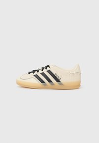 Vit sneaker med svarta mockastripes, strukturerad ovandel, krämfärgade accenter och gummisula; har svarta snörningar och branding.
