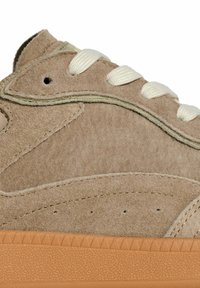 Primo piano di una sneaker in suede beige con lacci bianchi e suola in gomma leggera marrone texturizzata, evidenziando le cuciture e i fori di ventilazione.