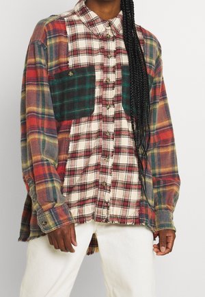 Persoon draagt een oversized ruitjesoverhemd met verschillende kleuren en knoopsluiting aan de voorkant, gecombineerd met een wit broek.