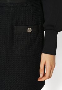 Zwarte geweven rok met een gestructureerde stof, een voorzak met een detail van een zwart lint en een accenten van een metalen knoop.