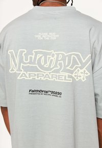 Světle šedé tričko s výrazným bílým nápisem "Multiply Apparel" na zadní straně, s pokyny k údržbě a detaily o značce v černé barvě ve spodní části.