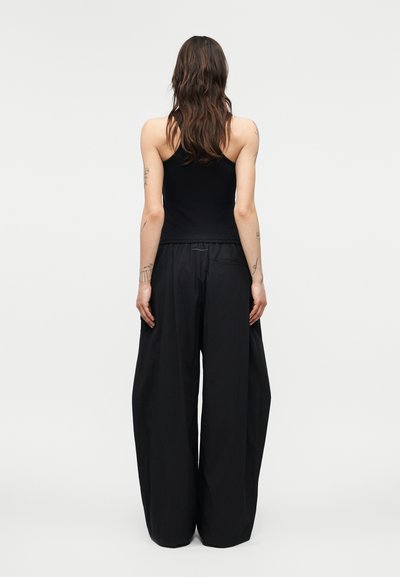 Mujer con cabello largo y oscuro, vistiendo una camiseta negra sin mangas y pantalones negros de pierna ancha, de pie de espaldas sobre un fondo liso.