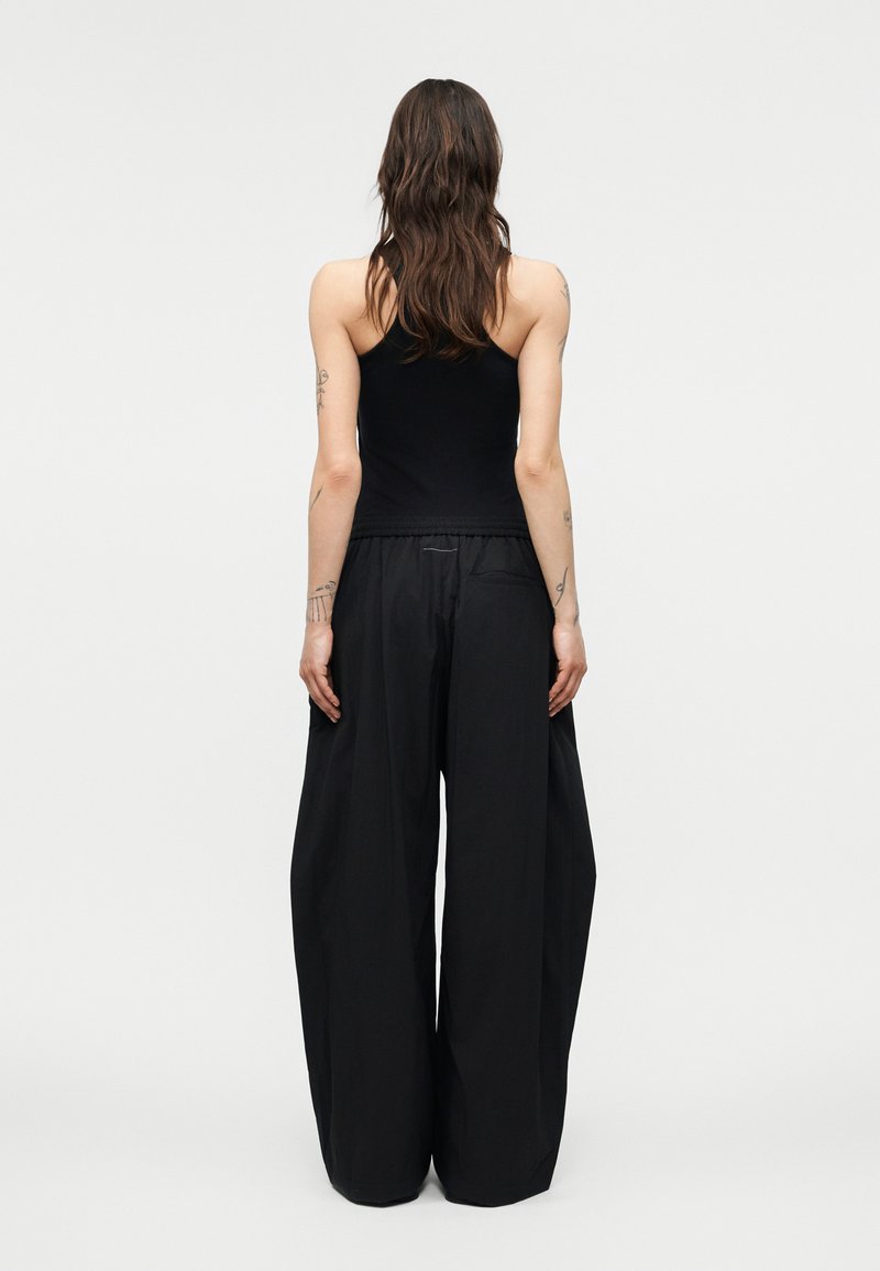 Femme aux longs cheveux foncés portant un haut noir sans manches et un pantalon noir ample, debout de dos sur un fond uni.