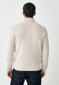 Pull léger beige clair avec un col montant, des poignets à côtes et un ourlet, porté sur un jean en denim foncé. Le tissu semble doux et texturé.