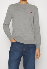 Szary dziergany sweter z okrągłym dekoltem i długimi rękawami. Na piersi ma małe czerwone logo. Połączony z ciemnoniebieskimi dżinsami.