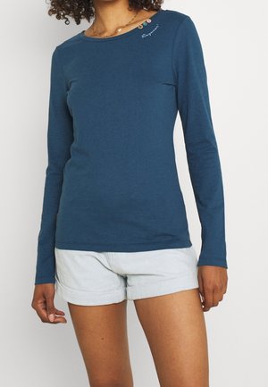 Femme portant une chemise bleue à manches longues avec trois petits boutons et un texte blanc près du col, associée à un short en jean clair.