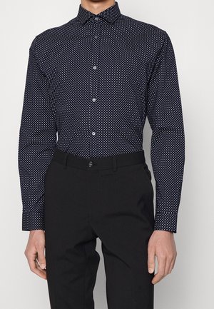 Muž nosící černou košili se vzorem bílých puntíků, zapnutou na knoflíky, a černé elegantní kalhoty, stojící před jednobarevným pozadím.