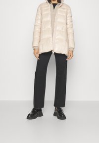 Osoba w jasnobeżowej kurtce puchowej, beżowym swetrze na zamek, czarnych spodniach o prostym kroju i czarnych, masywnych skórzanych butach stoi na jednolitym tle.