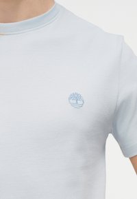 Ανοιχτό μπλε κοντομάνικο μπλουζάκι με στρογγυλή λαιμόκοψη, που φοριέται από ένα άτομο, με ένα μικρό κεντημένο λογότυπο δέντρου στο αριστερό στήθος.