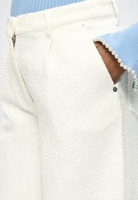 Main dans la poche d'un pantalon blanc texturé associée à une manche de pull bleu clair ornée d'une bordure de perles.