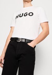 Mężczyzna w białej koszulce z napisem "HUGO" czarnymi literami, czarnym pasku i czarnych spodniach, z ręką częściowo w kieszeni, na jednolitym tle.