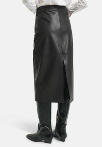 Zwarte leren midi-rok met een split aan de achterkant, aangevuld met strakke zwarte glanzende laarzen. Gladde textuur met minimale stikseldetails.